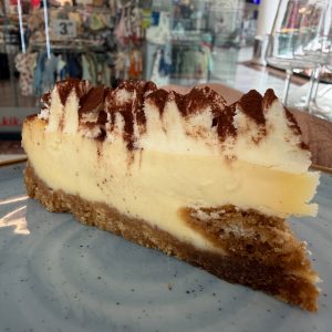 Slice of vanilla cheesecake with cocoa powder on top, served on a plate in a busy shop/mafe cafe background.