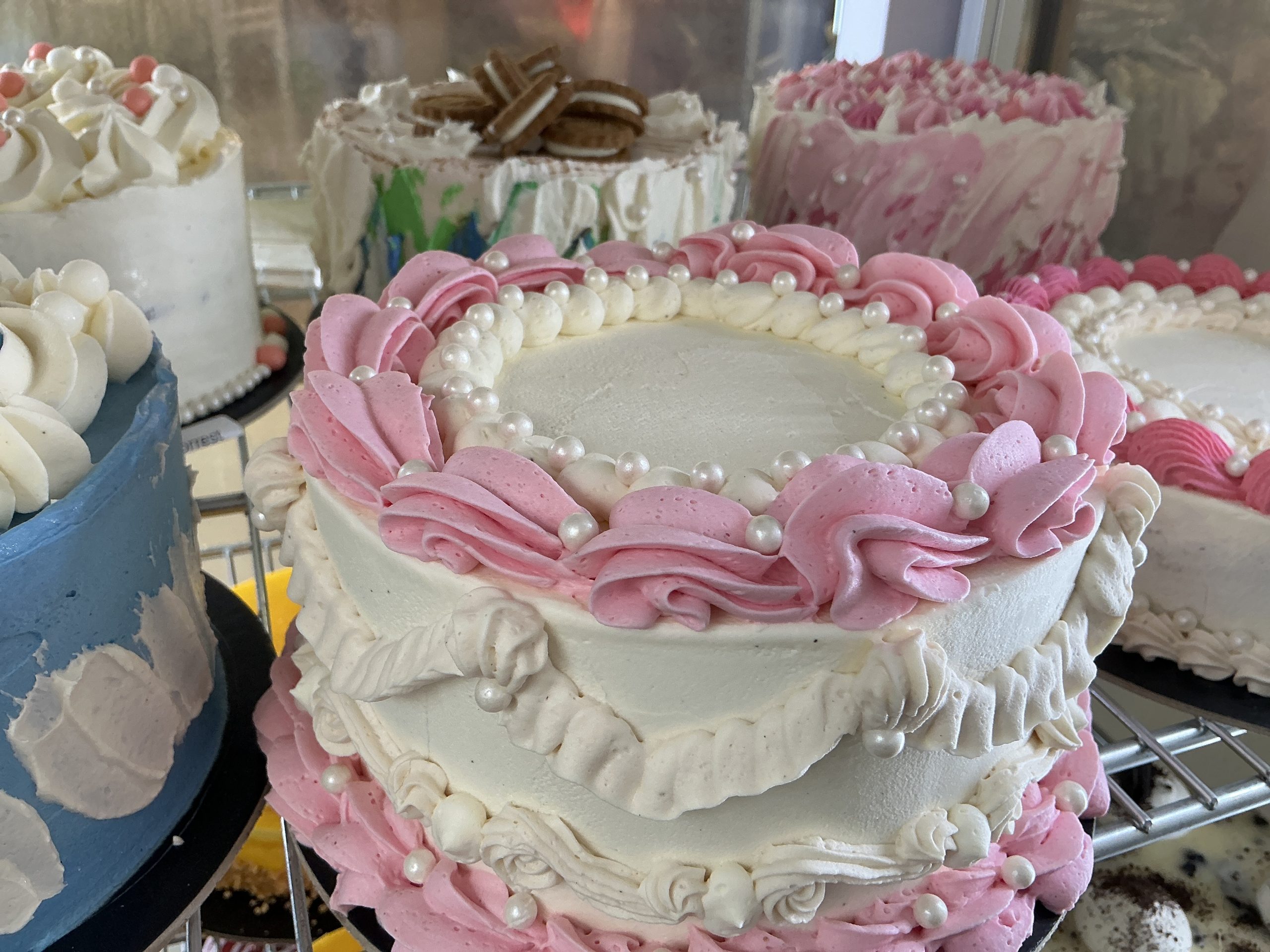 Close-up of pastel pink and white frosted cake with pearl decorations and piped rosettes on a bakery display stand.