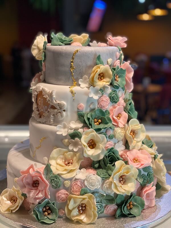 Three-tier wedding cake with white fondant and a cascade of pastel sugar flowers in cream, pink, mint, and gold accents.
