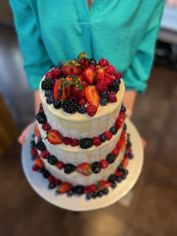 Three-tier white frosted cake decorated with strawberries, raspberries, blueberries and blackberries on top and around the tiers, held by a person in a teal shirt.