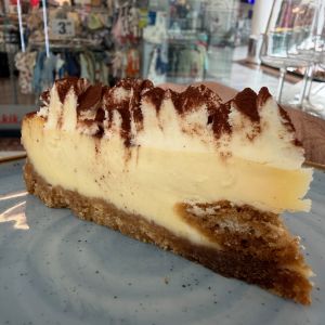 Slice of vanilla cheesecake with cocoa powder on top, served on a plate in a busy shop/mafe cafe background.